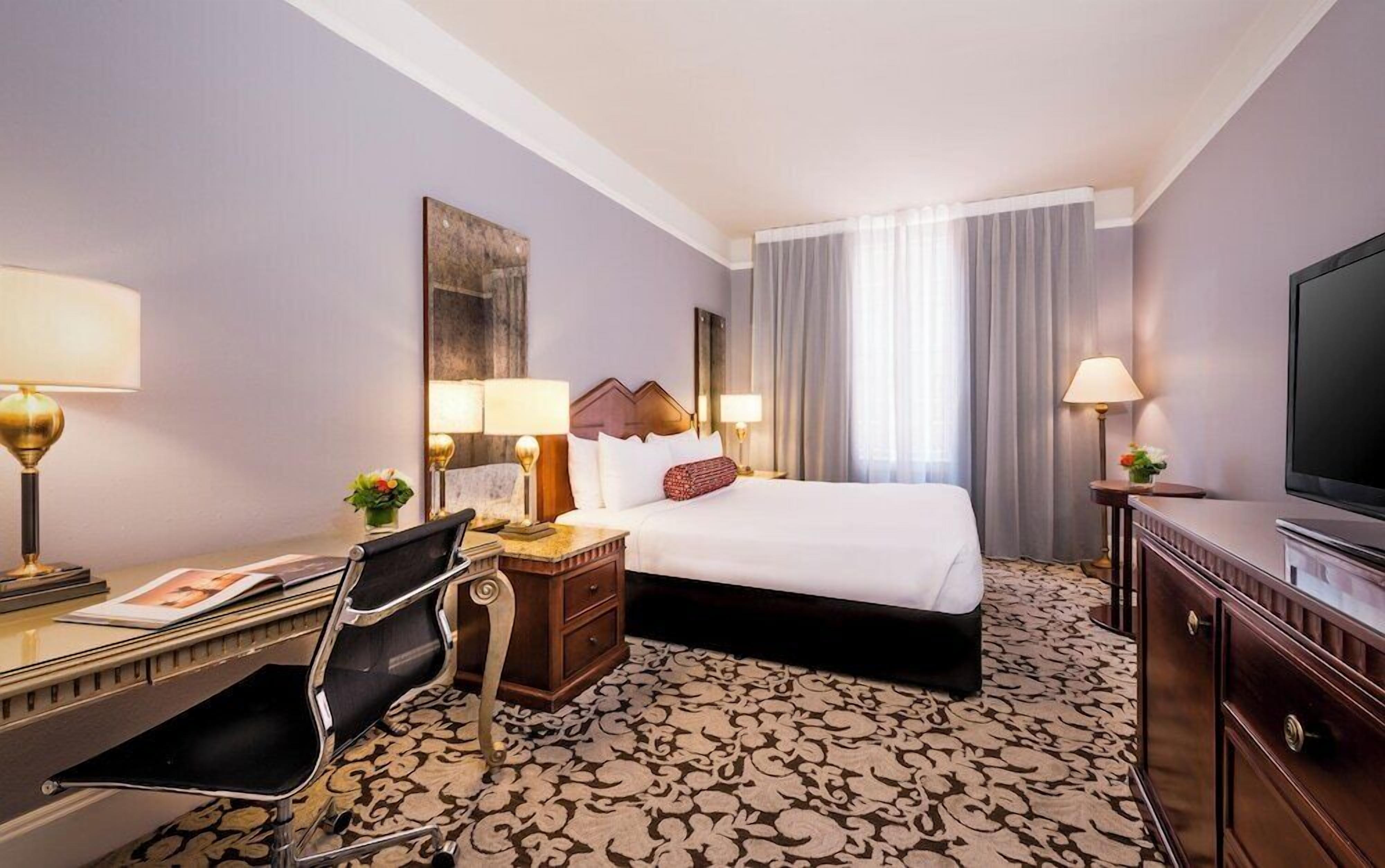 A hotel room with a bed, desk, chair, TV, and a patterned carpet.