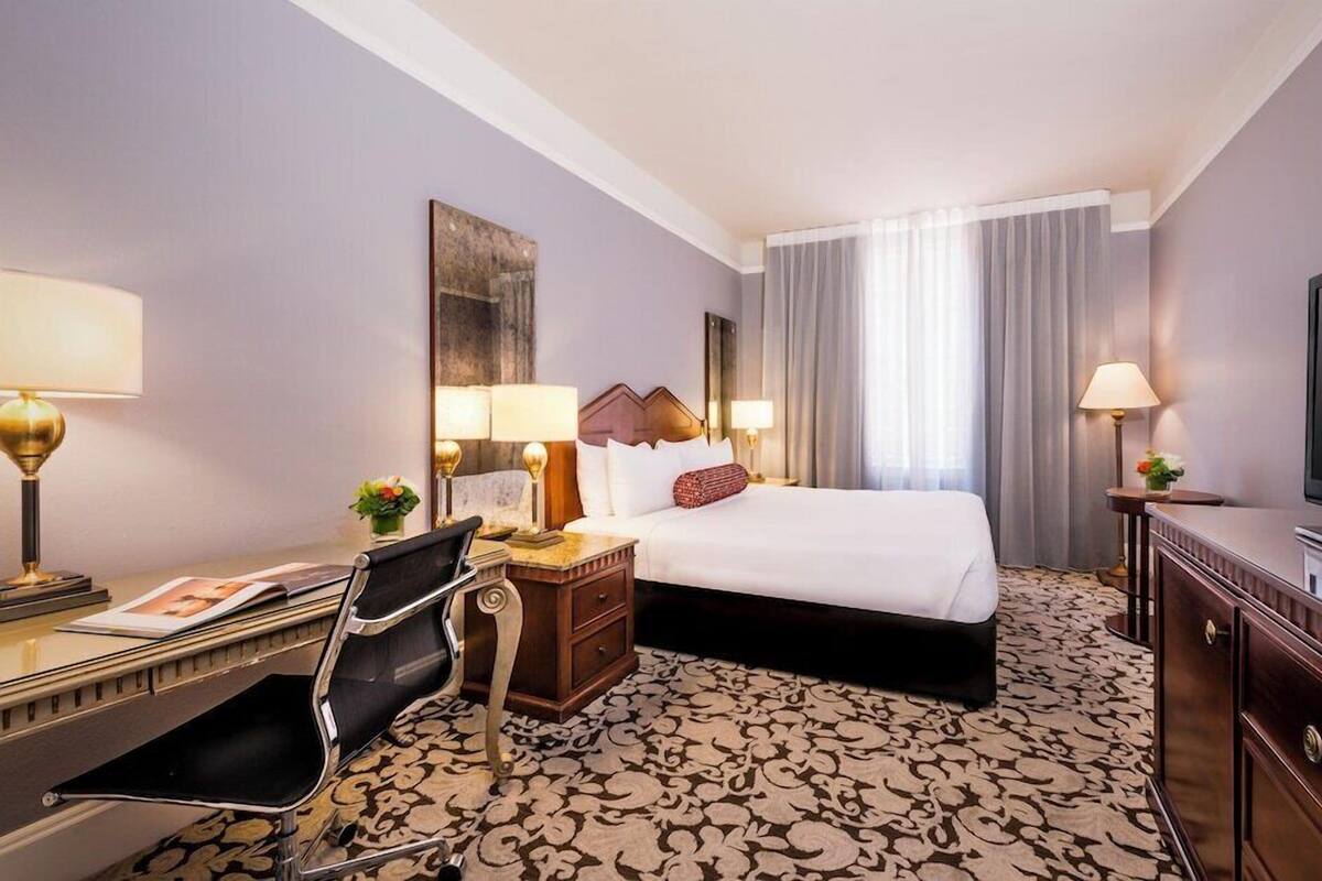 A hotel room with a bed, desk, chair, TV, and a patterned carpet.