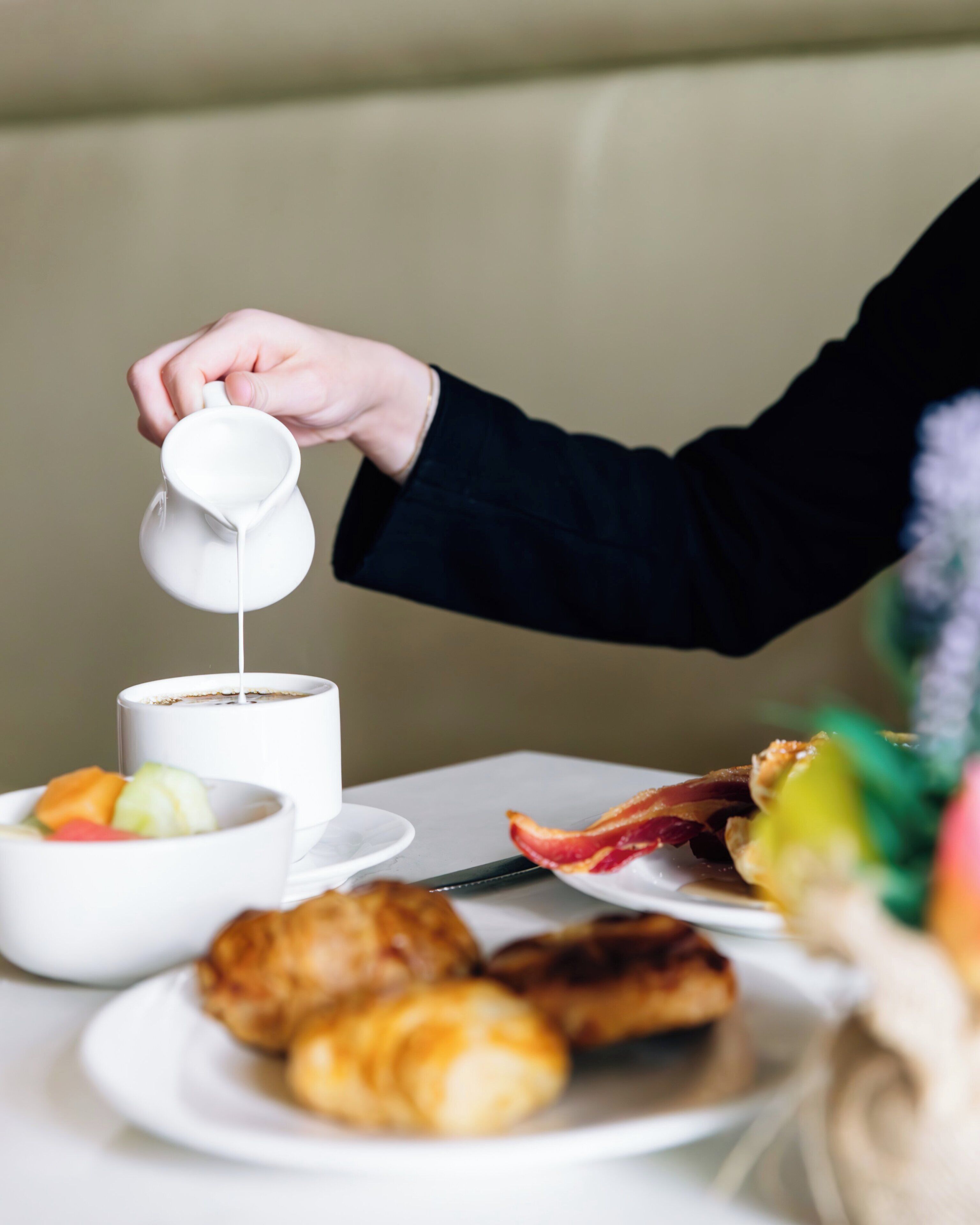 Una mano vertiendo leche en una taza de café, con croissants y fruta sobre la mesa.