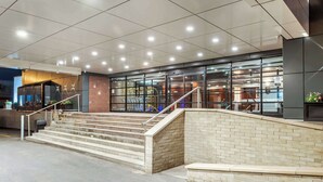 Ein moderner Gebäudeeingang mit einer Treppe, die zu einer Glastür führt, einer Ziegelwand und einem Metallgeländer.