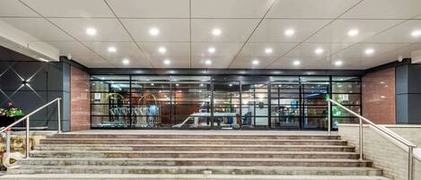A modern building entrance with a glass facade, a staircase leading up to the entrance, and a metal railing on the right side.