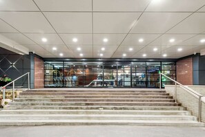 A modern building entrance with a glass facade, a staircase leading up to the entrance, and a metal railing on the right side.