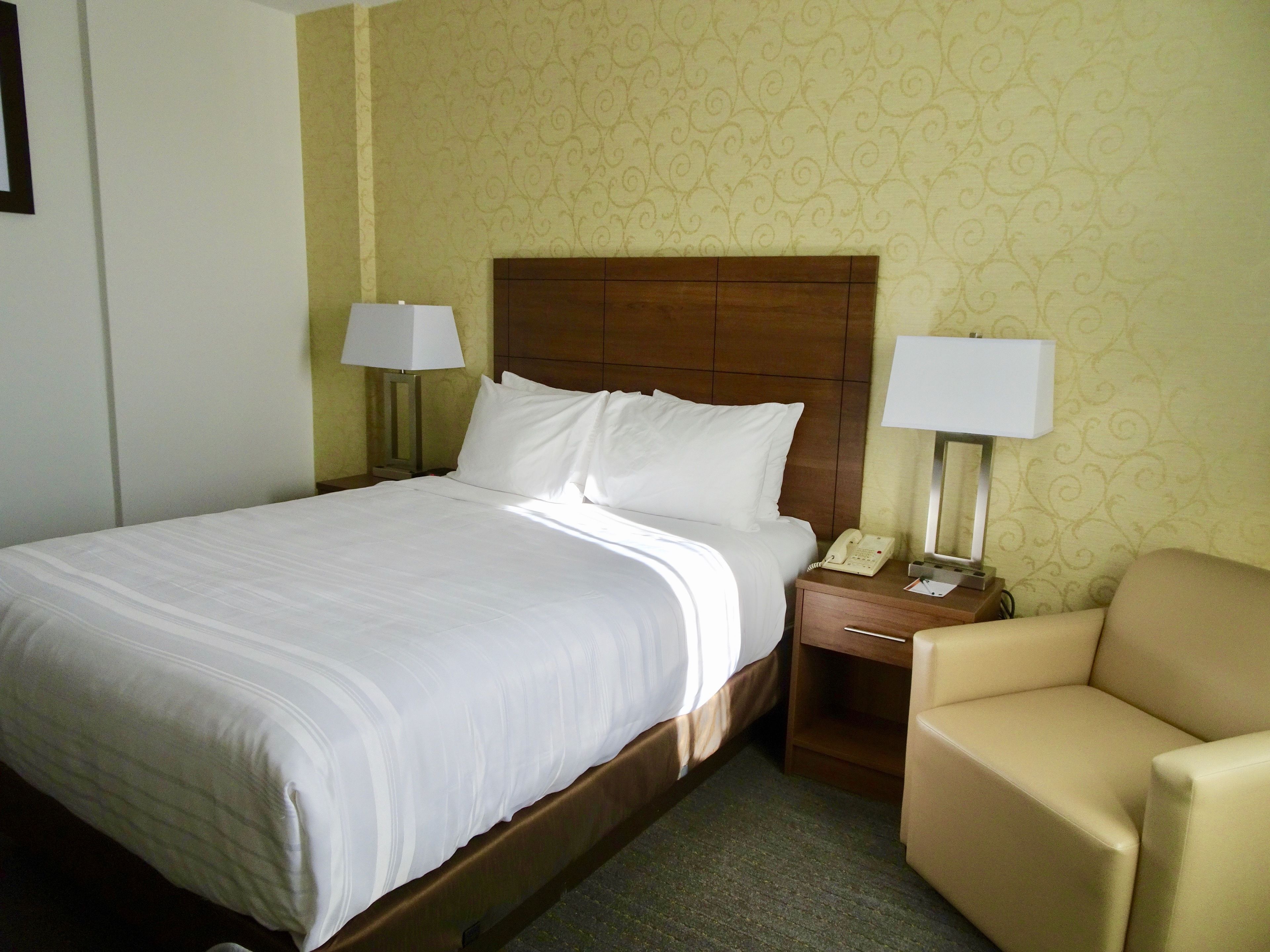 Une chambre d’hôtel avec un grand lit, deux lampes de chevet, une tête de lit en bois, un téléphone sur la table de chevet et un fauteuil beige.