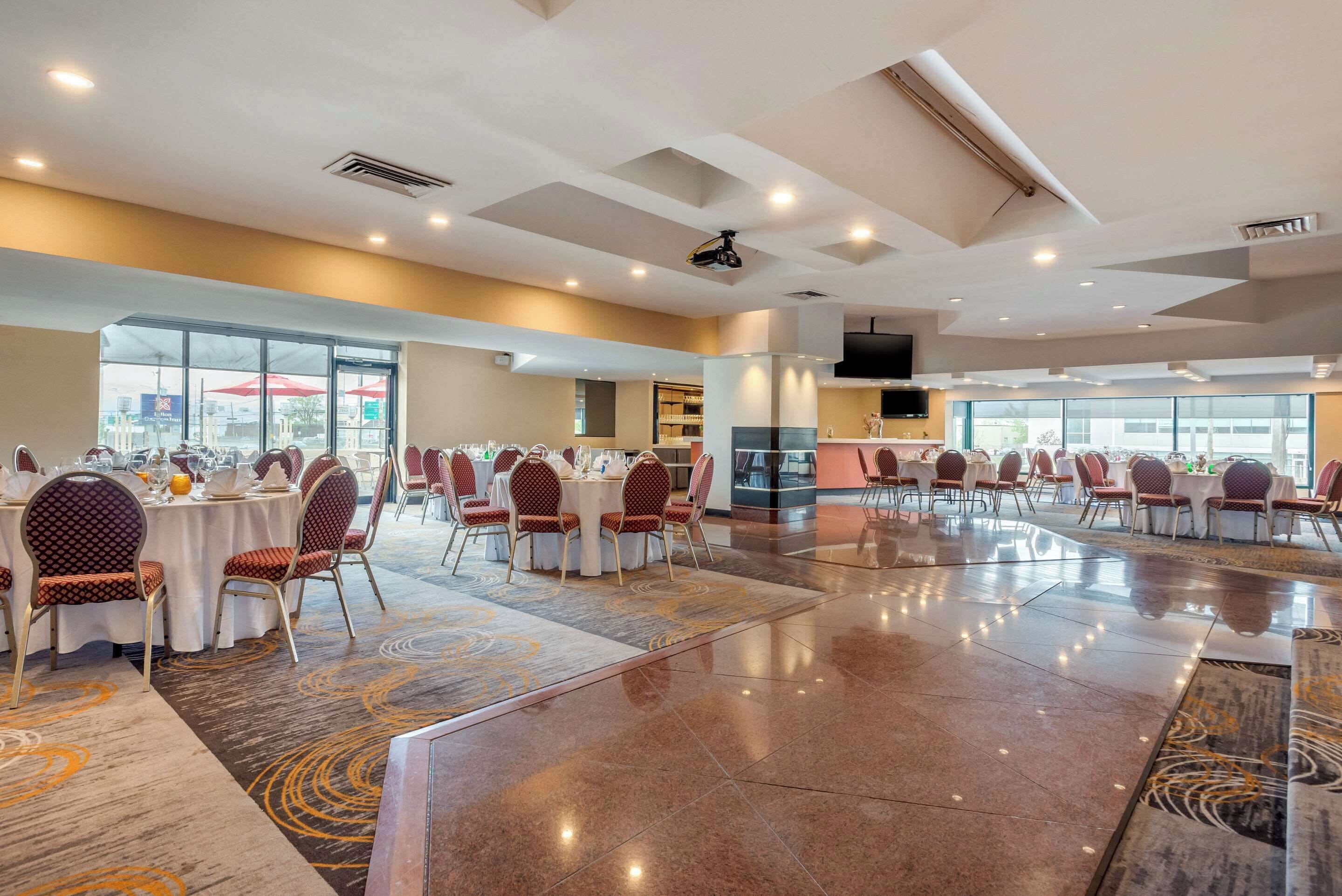 A spacious banquet hall with round tables and chairs, large windows, and a modern ceiling design.