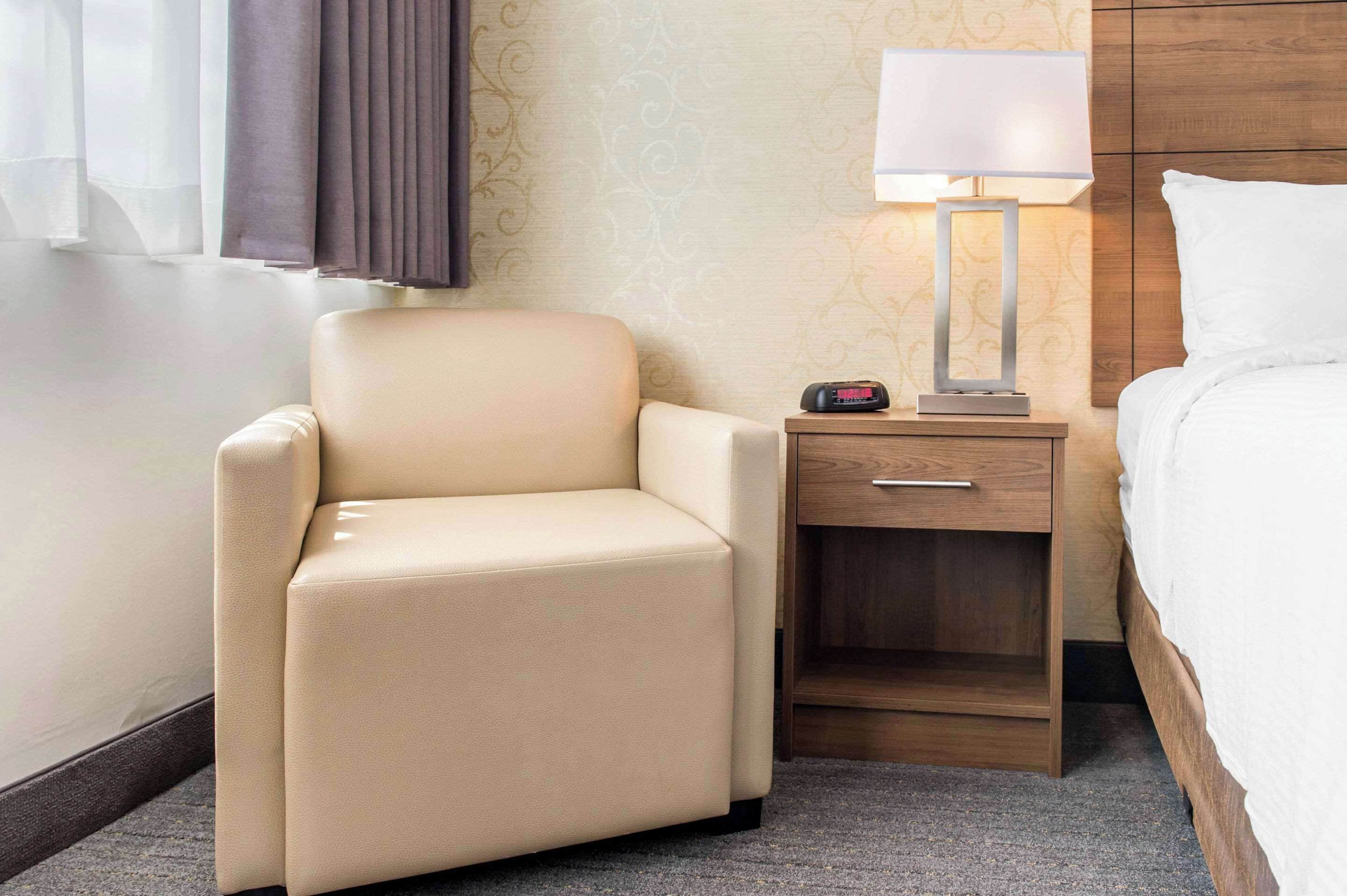 Une chambre d’hôtel avec un fauteuil beige, une table de chevet avec une lampe et un lit avec une literie blanche.