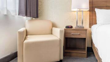 Une chambre d’hôtel avec un fauteuil beige, une table de chevet avec une lampe et un lit avec une literie blanche.