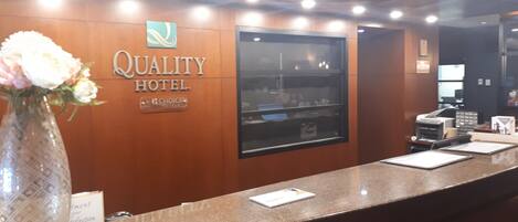 A reception desk at the Quality Hotel with a logo, a glass display case, and a vase with flowers.