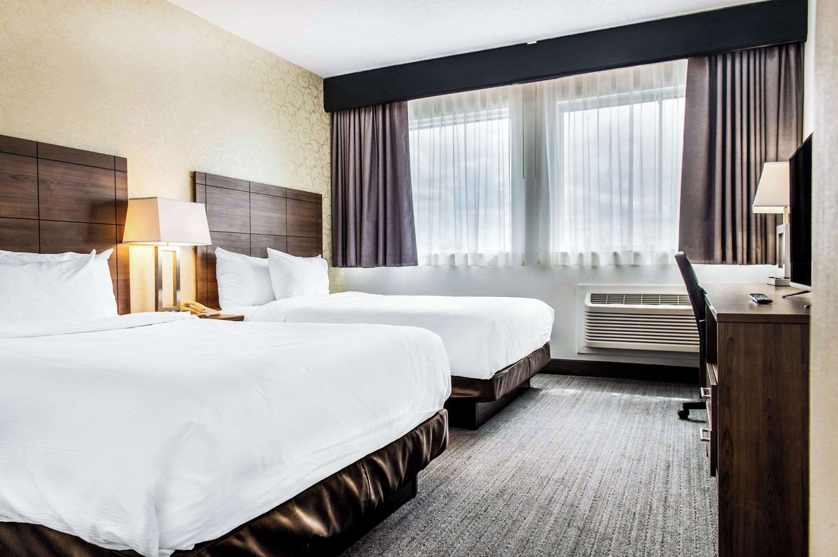 A hotel room with two beds, a desk, a chair, a lamp, and a large window with curtains.