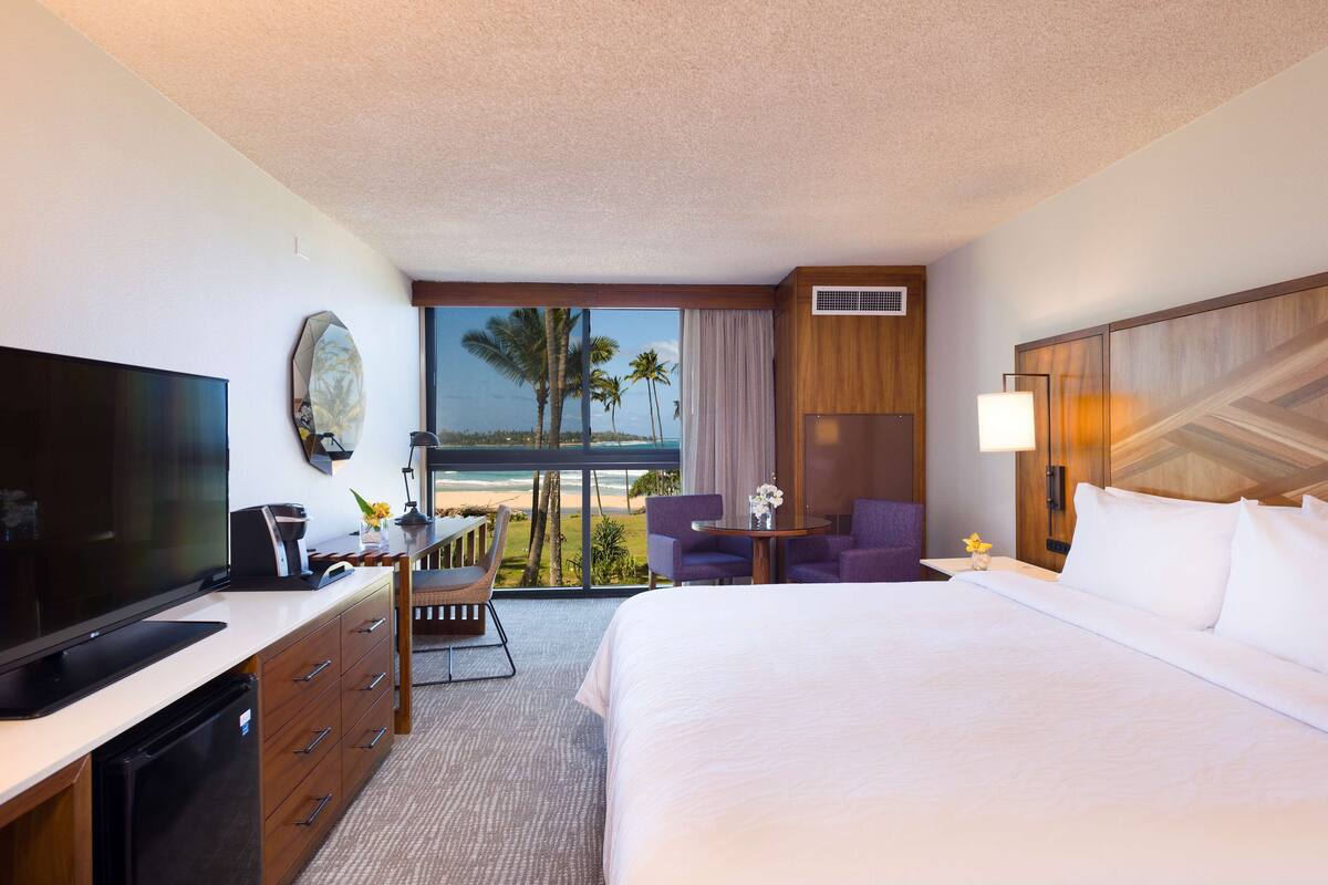 A hotel room with a bed, a desk with a TV, a chair, a small table with a lamp, and a view of the beach.