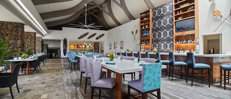 Un vaste espace de restauration intérieur, doté de nombreuses tables et chaises, d’un mur en pierre, d’un bar avec des étagères et d’un plafond avec des poutres apparentes.