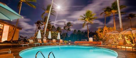 A swimming pool with lounge chairs and umbrellas, surrounded by palm trees and a building.