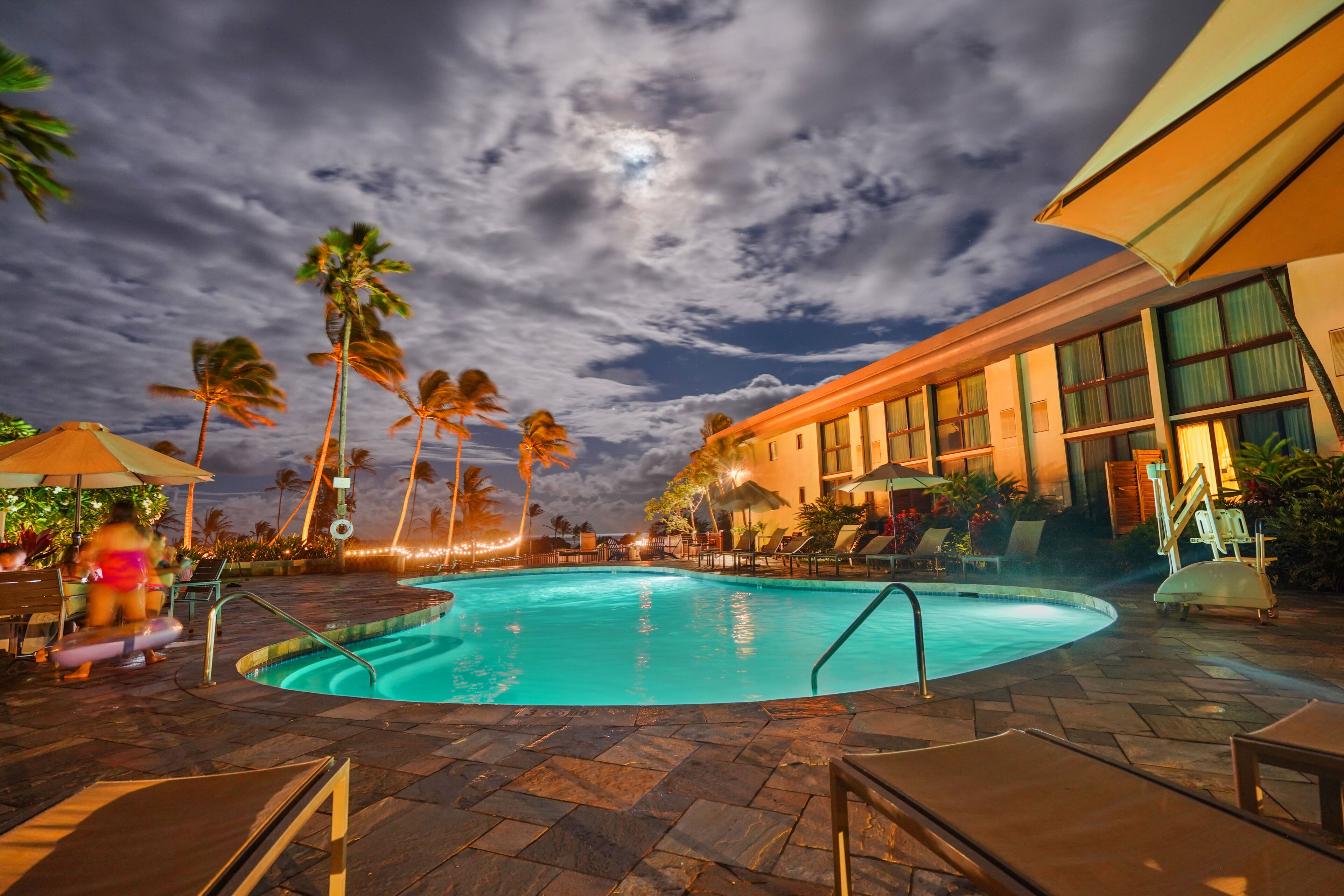 A pool area with lounge chairs and umbrellas, palm trees, and a building with large windows.
