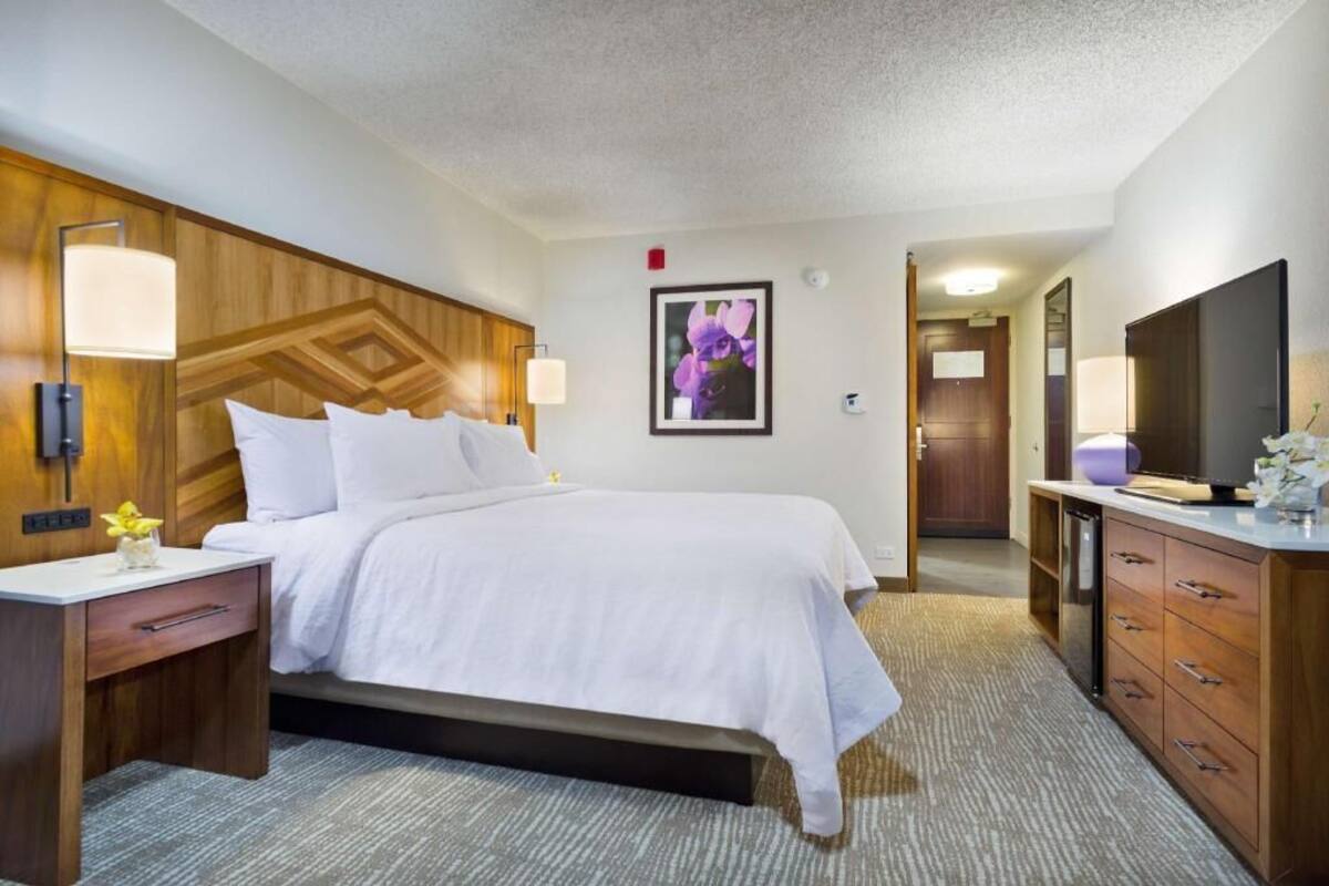 A hotel room with a bed, bedside tables, a TV, a mirror, and a framed picture on the wall.