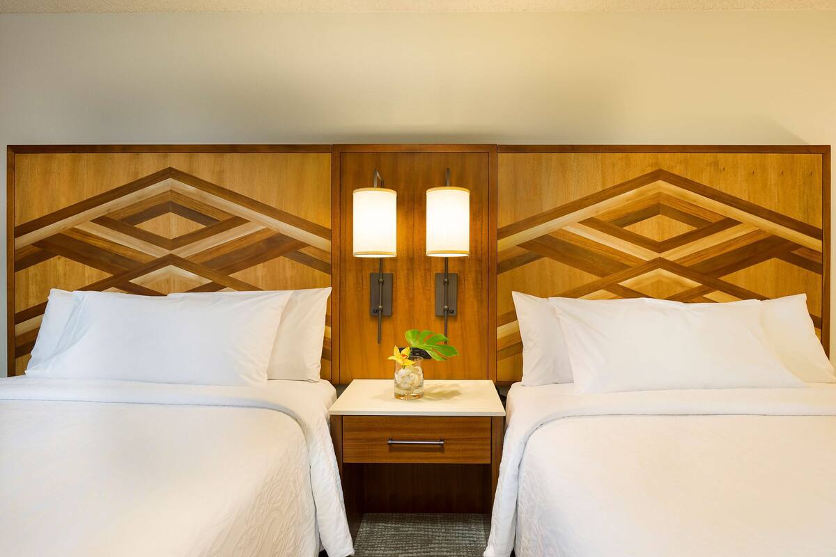 A hotel room with two beds, a wooden headboard with geometric patterns, a nightstand with two wall-mounted lamps, and a small vase with a plant.