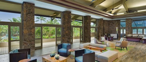 A spacious indoor area with stone pillars, comfortable seating, and large windows offering views of palm trees.