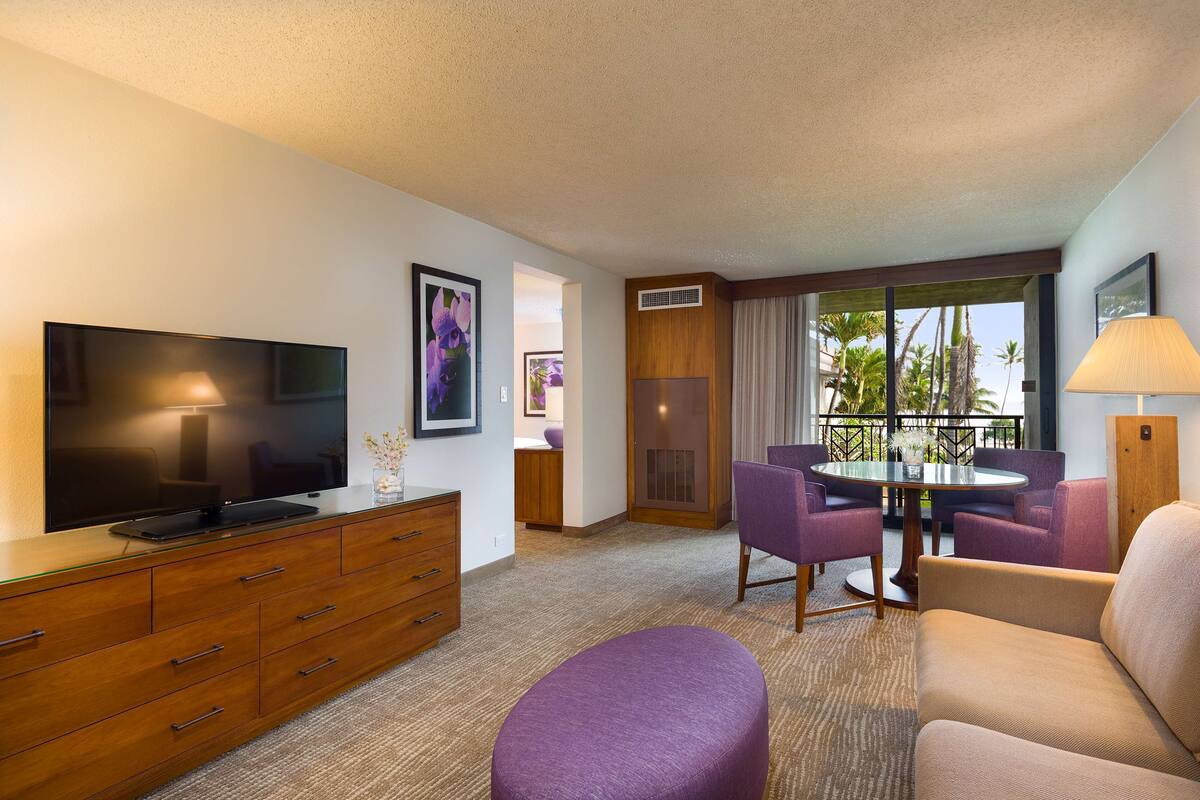 A hotel room with a flat-screen TV, a wooden dresser, a purple ottoman, a beige sofa, a round table with purple chairs, and a view of palm trees through a large window.