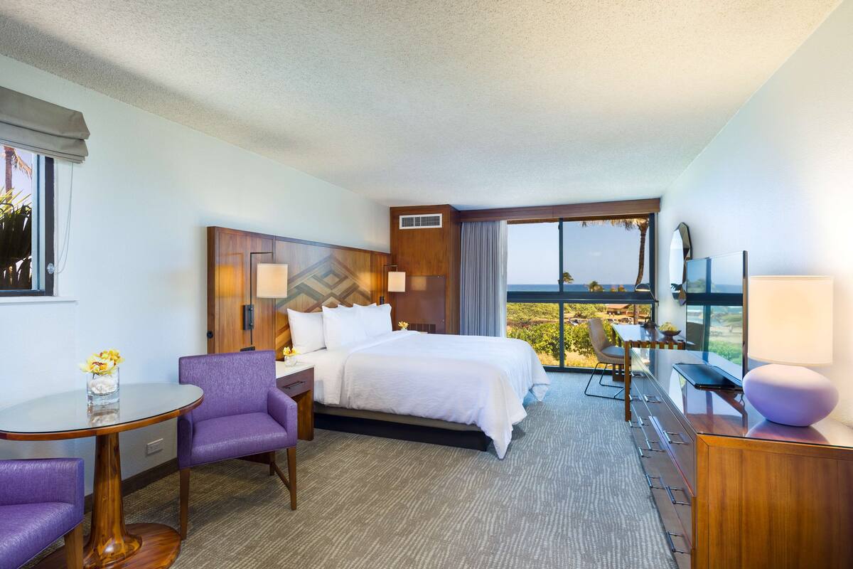 A hotel room with a bed, a desk, a chair, a small table, and a view of the outdoors.