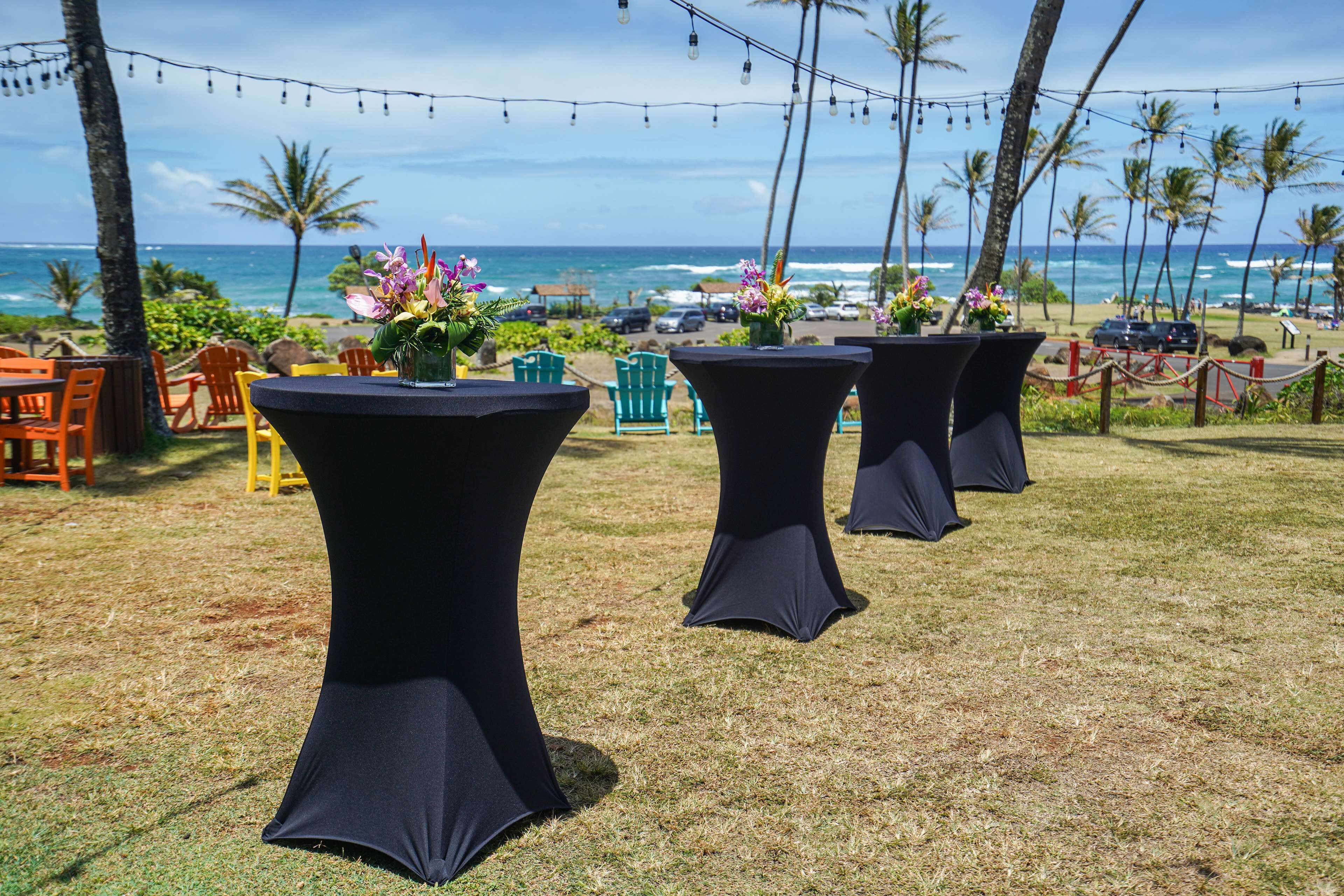 Outdoor tables with black tablecloths, chairs, and flower arrangements against a backdrop of the ocean and palm trees.