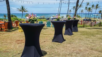 Des tables installées à l'extérieur, recouvertes de nappes noires, entourées de chaises et agrémentées de compositions florales, le tout se dressant sur fond d'océan et de palmiers.