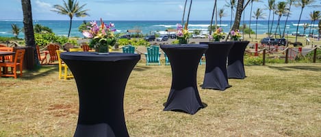 Outdoor tables with black tablecloths, chairs, and flower arrangements against a backdrop of the ocean and palm trees.