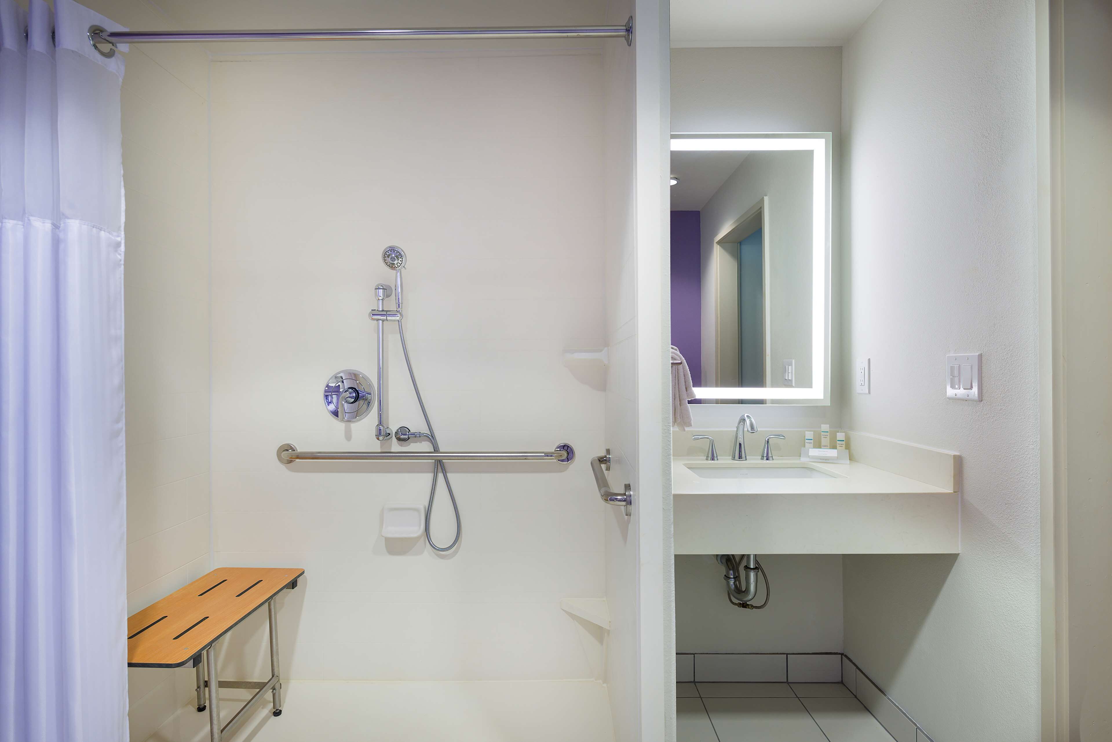 A bathroom with a shower, a grab bar, a wooden bench, a sink, and a mirror.