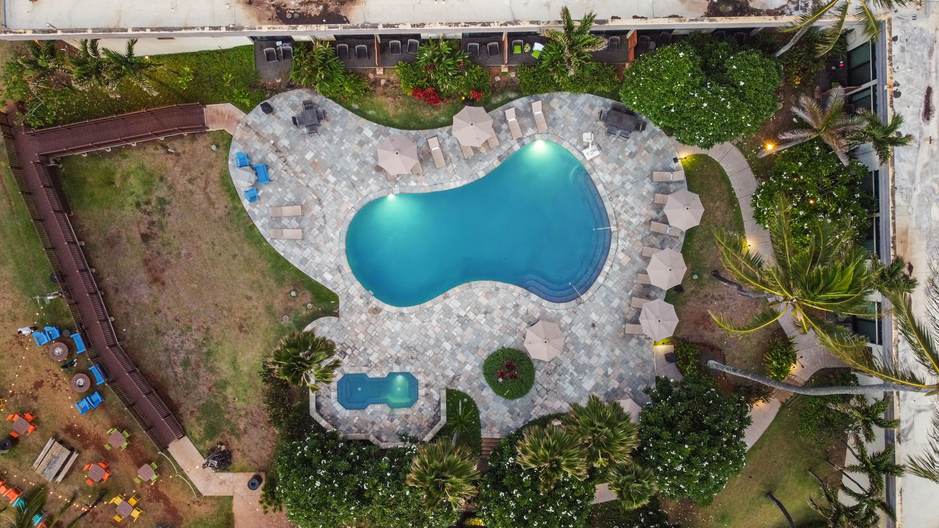 Vista aérea de un área de piscina con una piscina central grande, piscinas más pequeñas y zonas de descanso circundantes con mobiliario y sombrillas.
