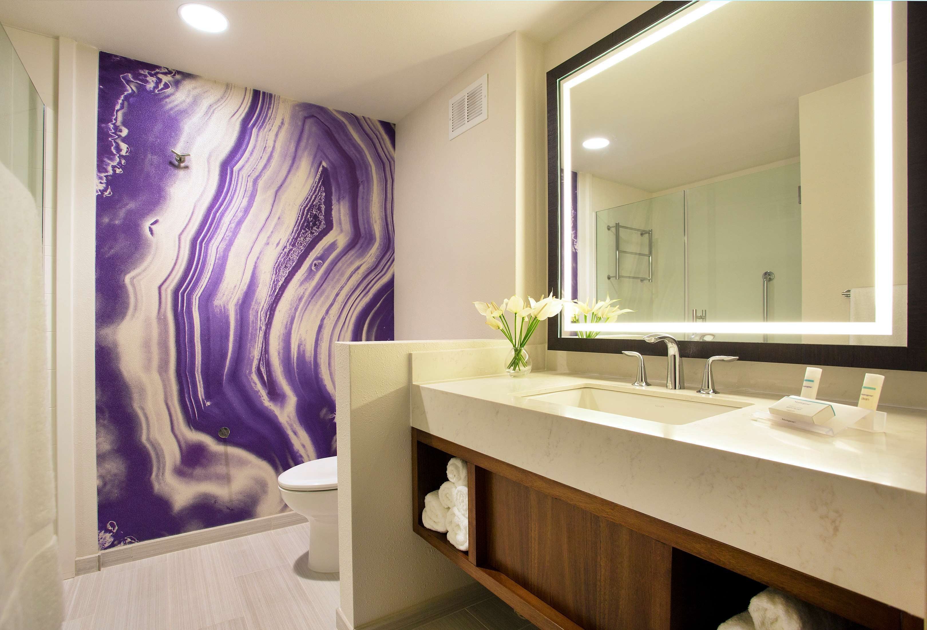 Un baño moderno con pared de mármol, un espejo grande, un lavamanos y un inodoro.