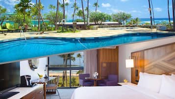A hotel room with a view of a swimming pool and beach, a desk with a coffee maker, and a bed with white linens.