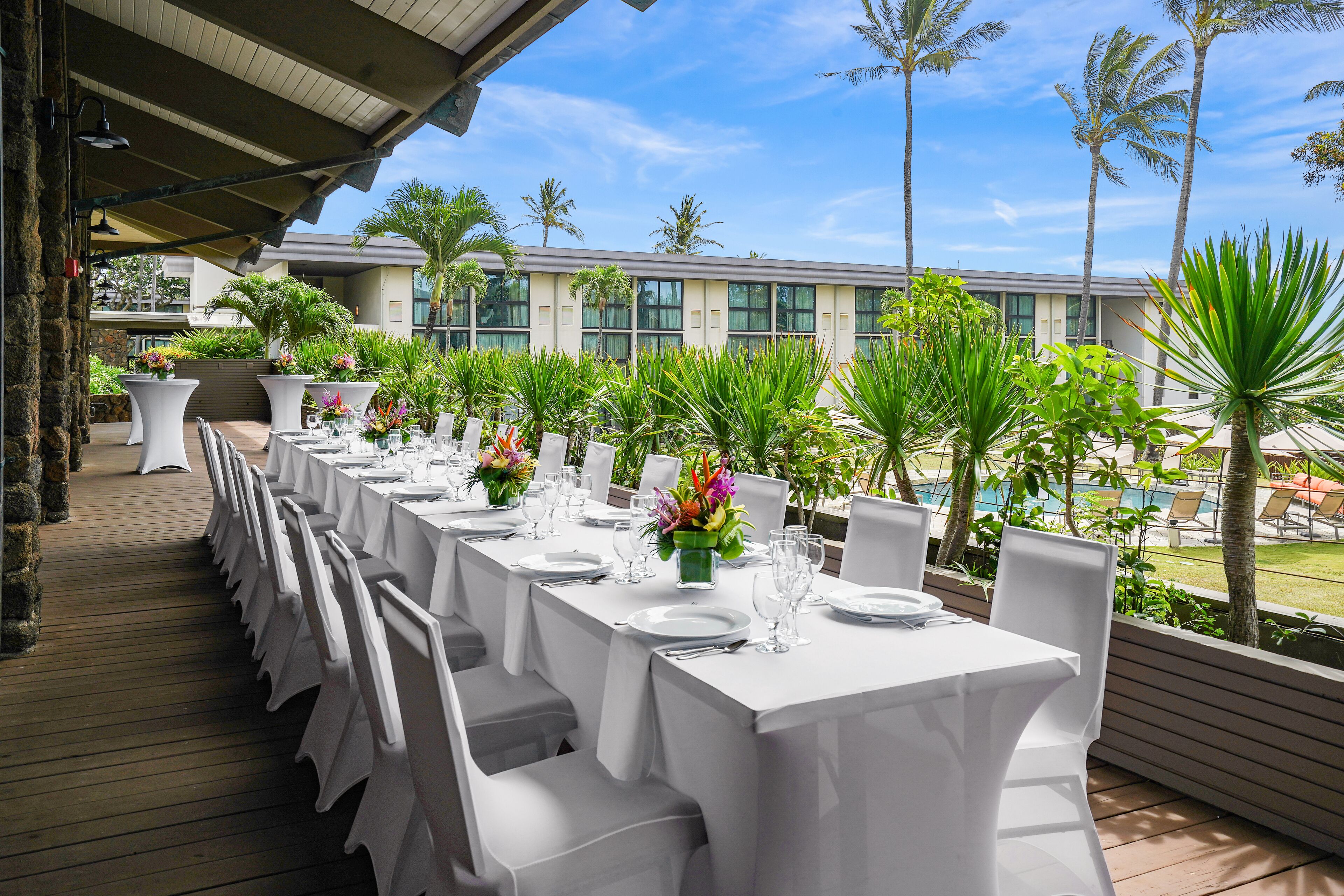 A long table set for dining with white tablecloths and chairs, surrounded by tropical plants and palm trees, with a pool and building in the background.
