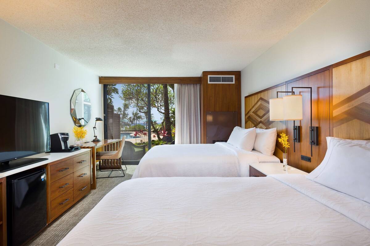A hotel room with two beds, a desk with a television, a minibar, and a view of the outdoors.