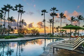 A poolside area with lounge chairs and umbrellas, palm trees, and a sunset view.