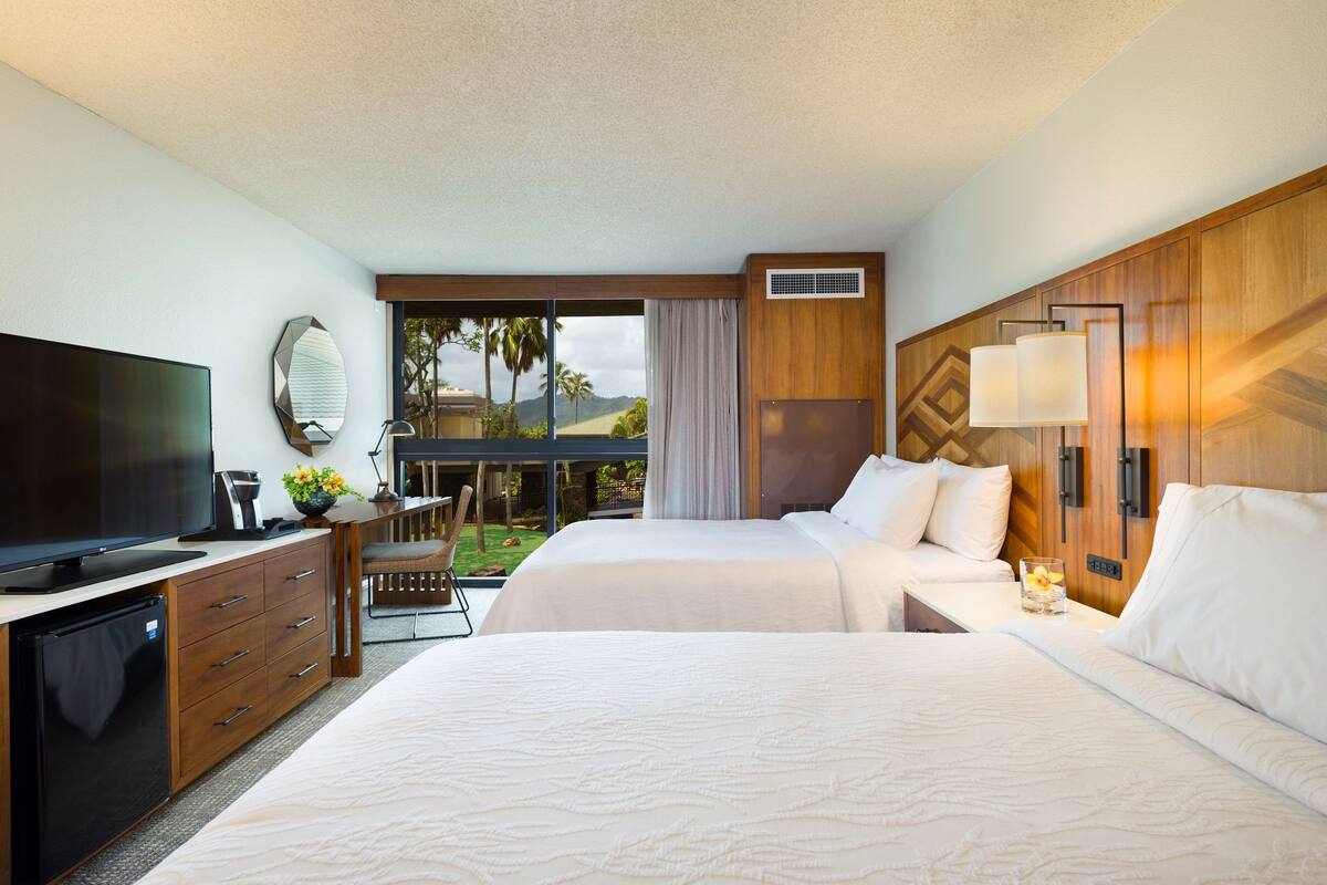 A hotel room with a TV, a desk with a lamp, two beds, a window with a view of palm trees, and a balcony.