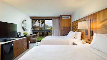 Une chambre d’hôtel équipée d’une télévision, d’un bureau avec une lampe, de deux lits, d’une fenêtre donnant sur des palmiers et d’un balcon.