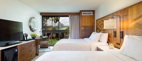Une chambre d’hôtel équipée d’une télévision, d’un bureau avec une lampe, de deux lits, d’une fenêtre donnant sur des palmiers et d’un balcon.