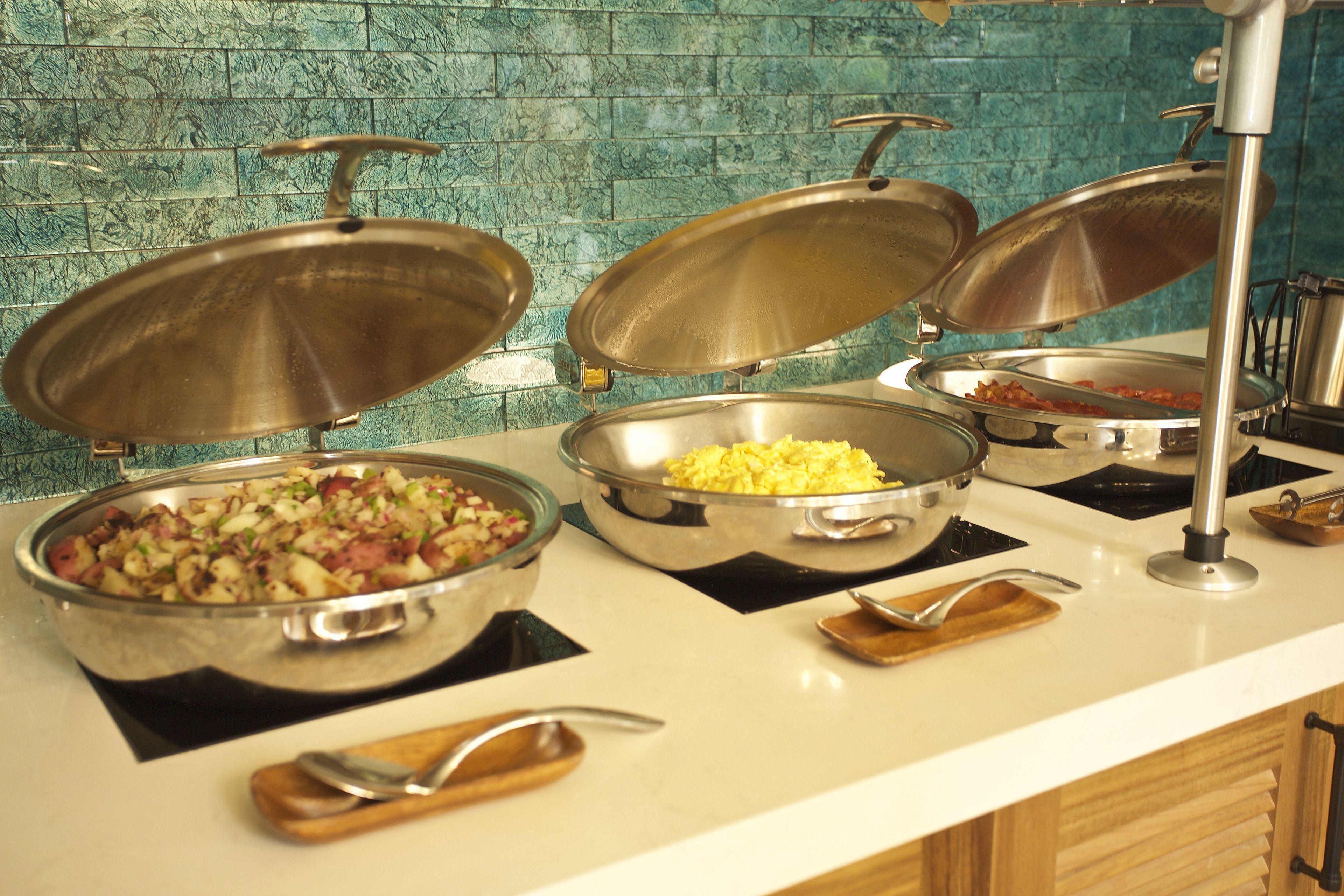 Ein reichhaltiges Buffet mit verschiedenen Speisen in Metallservierbehältern, einem Holzernst und einem Löffel.