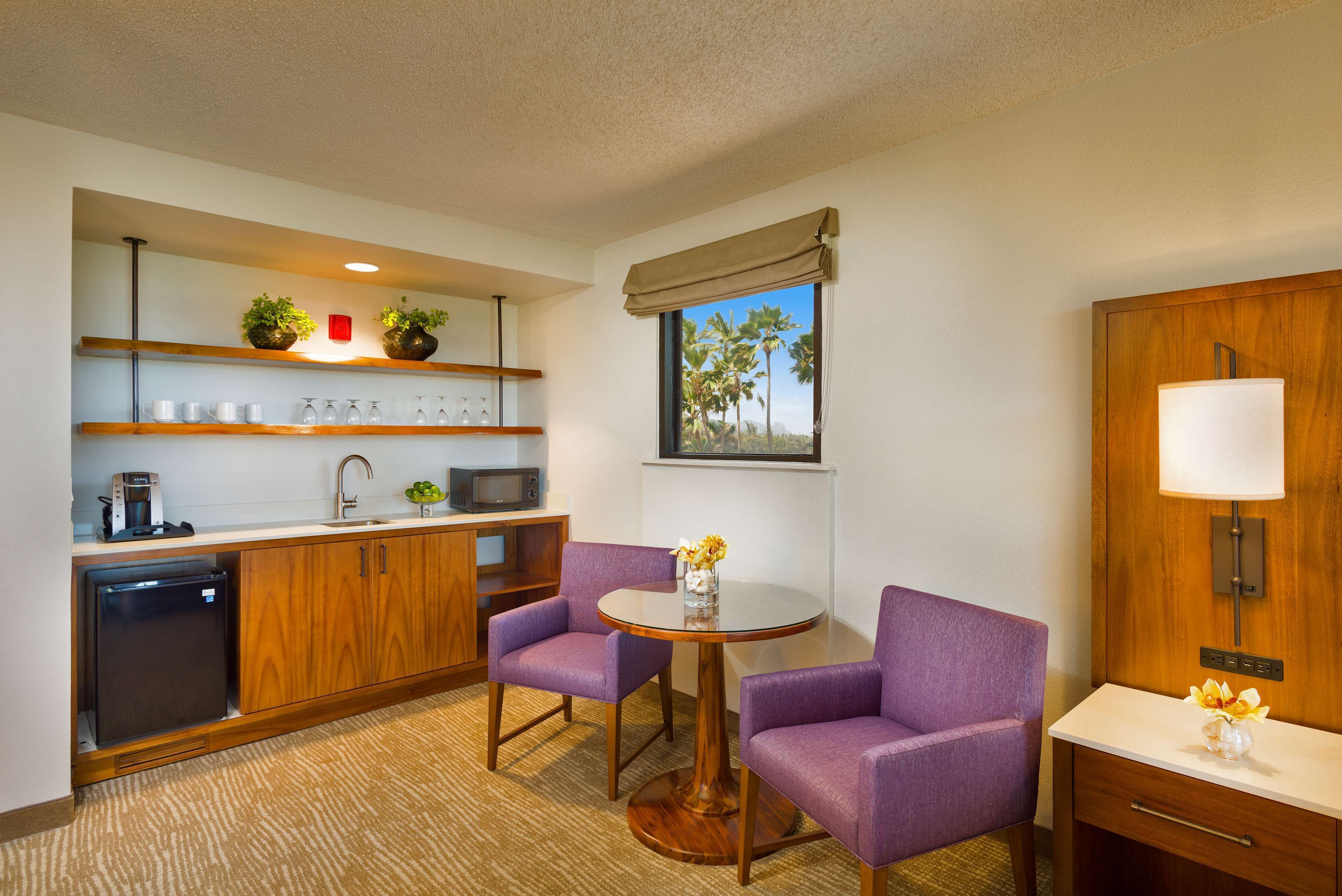 Una habitación de hotel compacta con kitchenette, dos sillones morados, una pequeña mesa redonda, un armario de madera, microondas, refrigerador, fregadero, ventana con vista a palmeras y una lámpara fijada a la pared.