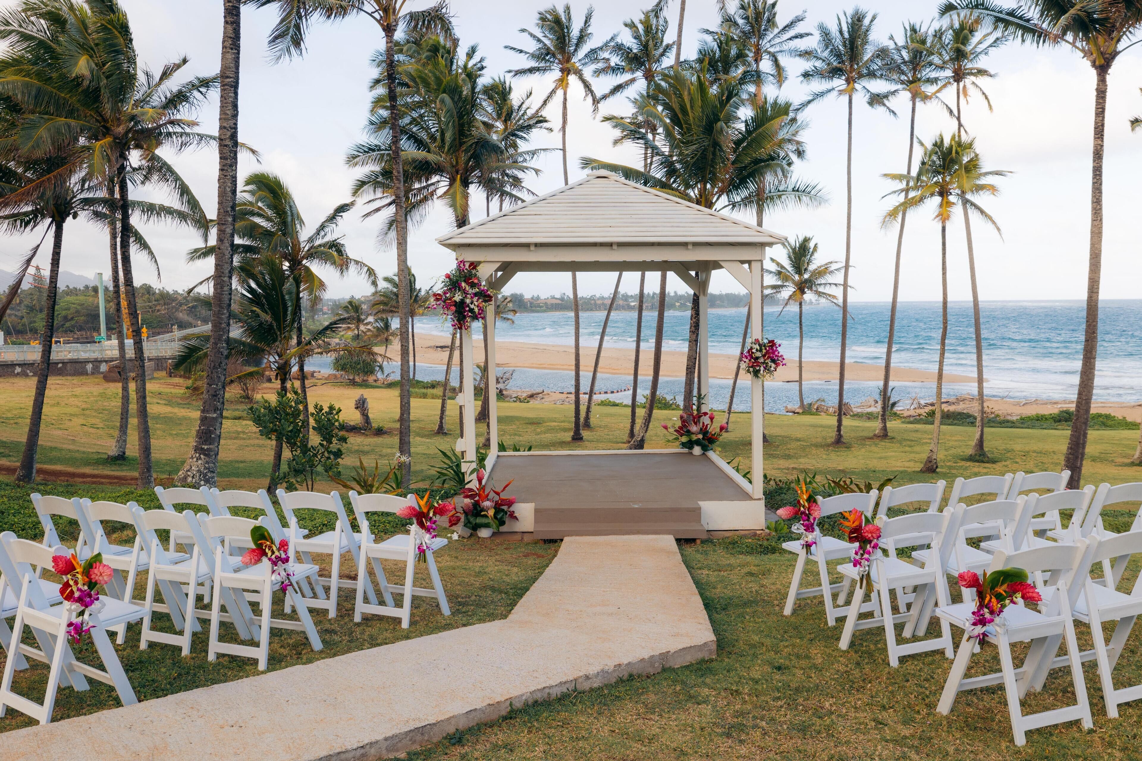 Un pabellón con sillas blancas y arreglos florales, con vista a una playa con palmeras.