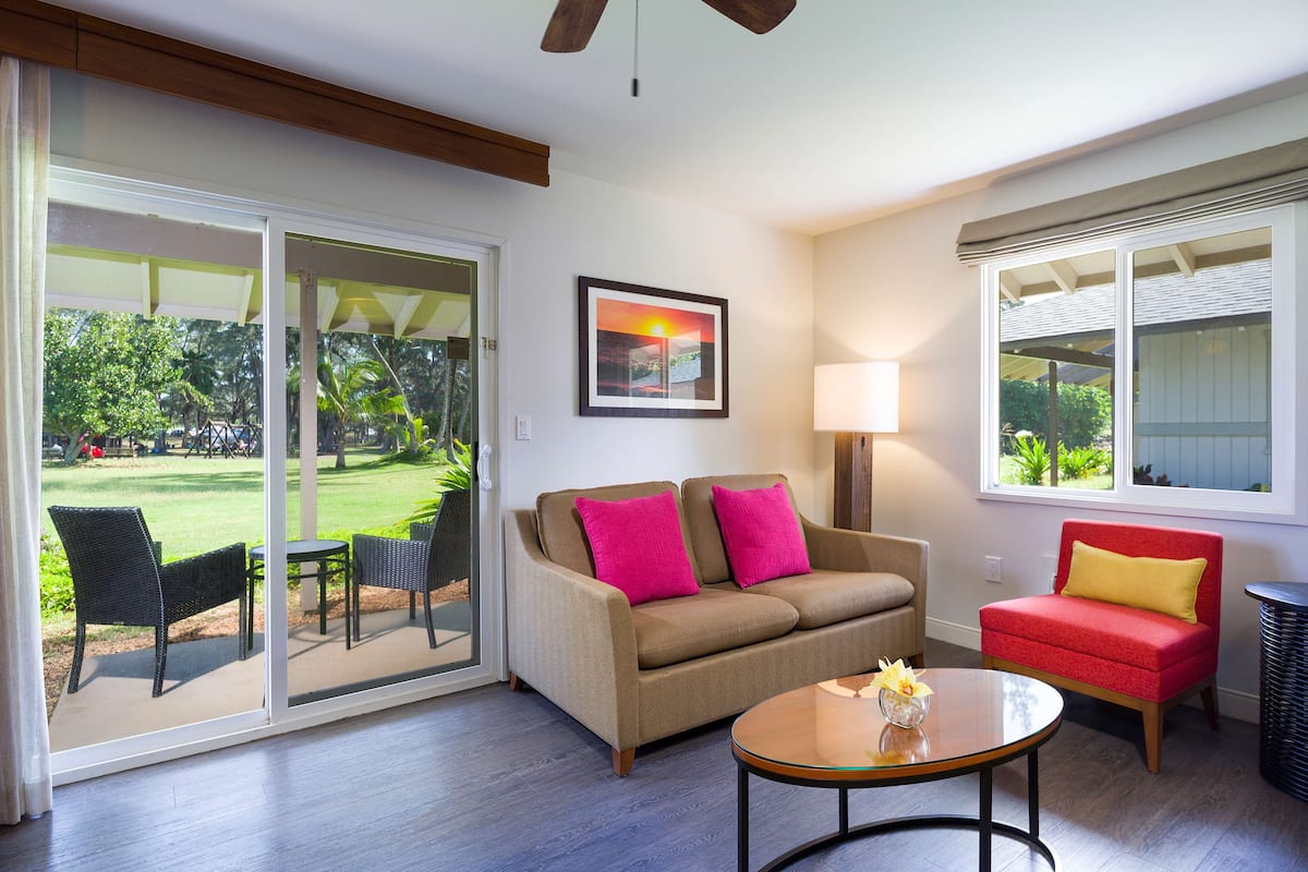 A living room with a sliding glass door leading to a patio, a beige sofa with pink cushions, a red armchair, a wooden coffee table, and a lamp.