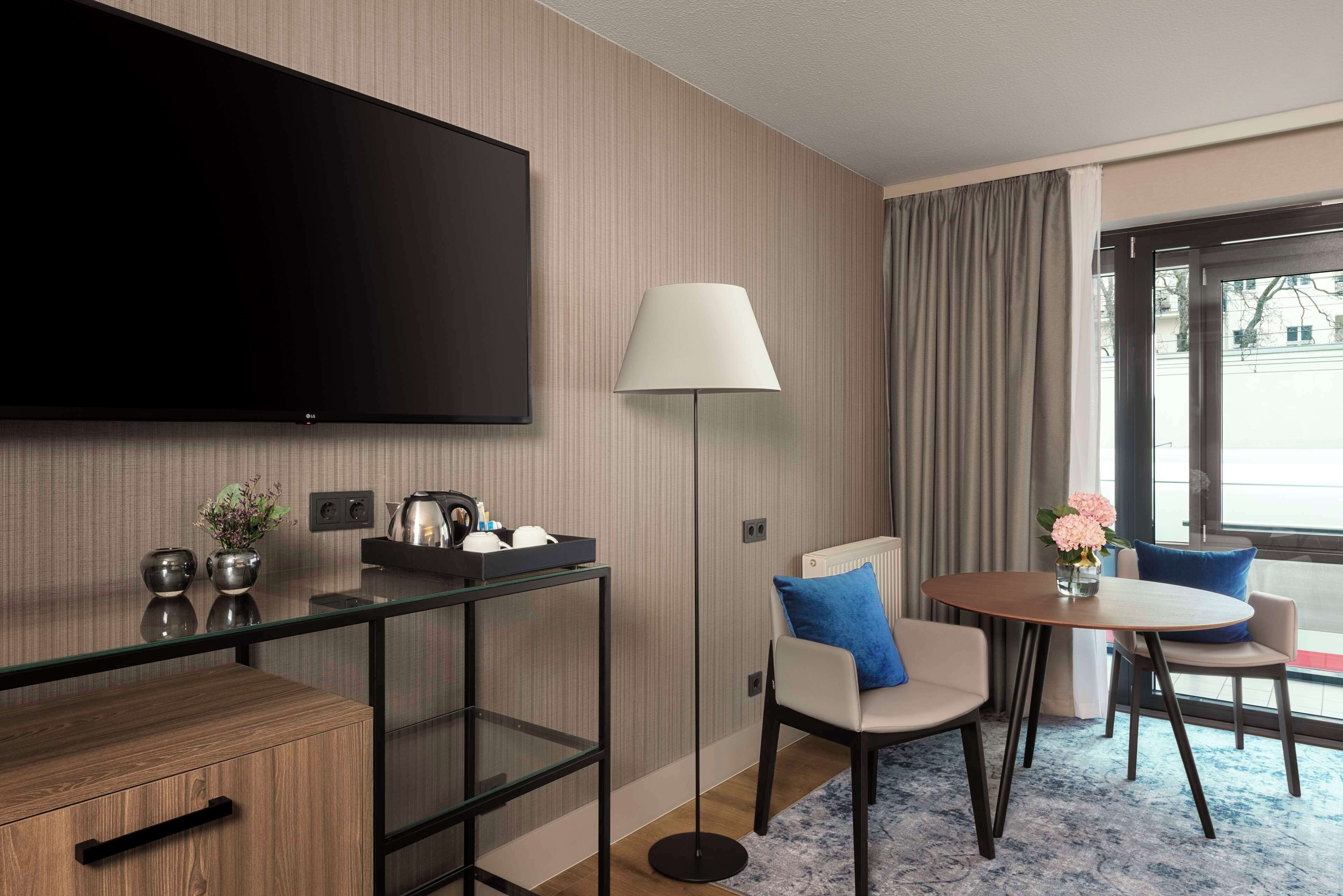 A modern hotel room with a flat-screen TV, a glass coffee table, a round dining table with chairs, a floor lamp, and a view of the outside through a large window.