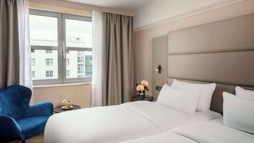 Quarto de hotel com uma cama grande, uma poltrona azul, uma mesinha de centro com um vaso de flores e uma janela com cortinas.