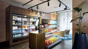 Un hall d’hôtel moderne doté d’un présentoir réfrigéré pour les boissons, d’étagères à collations, d’un comptoir de réception et d’une grande plante en pot.