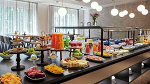 Un buffet proposant une grande variété de plats et de boissons, notamment des fruits, des fromages et des salades, dans un hôtel moderne doté de grandes fenêtres et d'un éclairage décoratif suspendu.