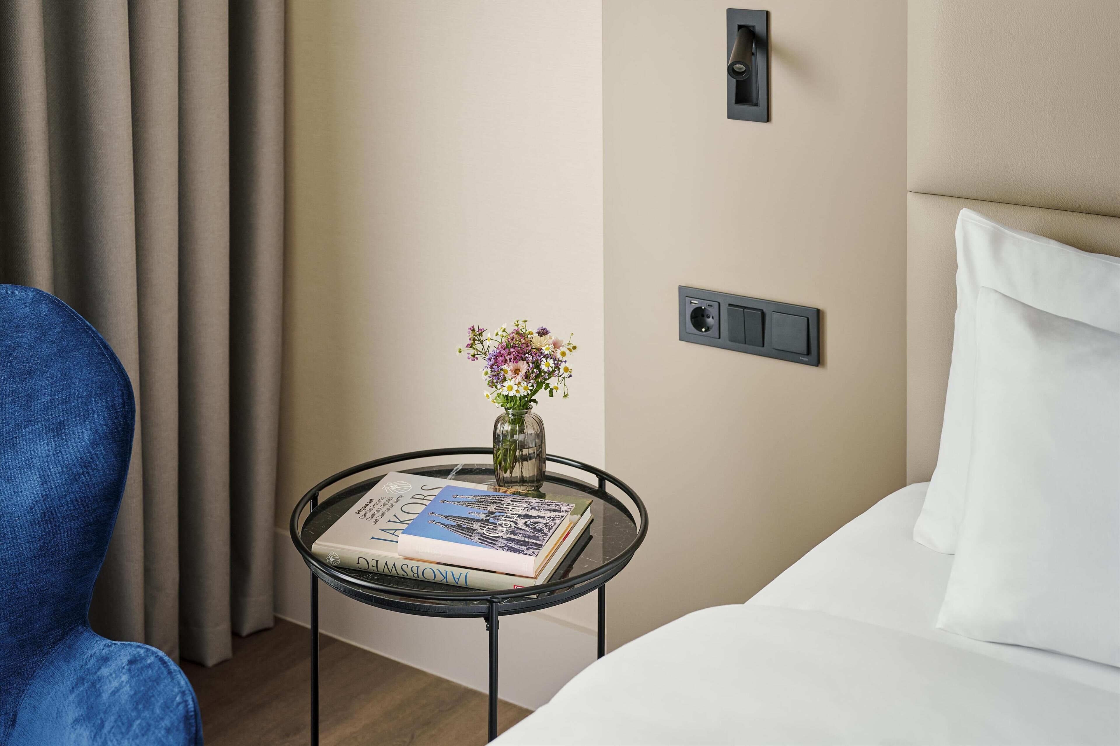 Une chambre d’hôtel avec un lit, une chaise bleue, une petite table avec un livre et un vase de fleurs, et une prise de courant au mur.