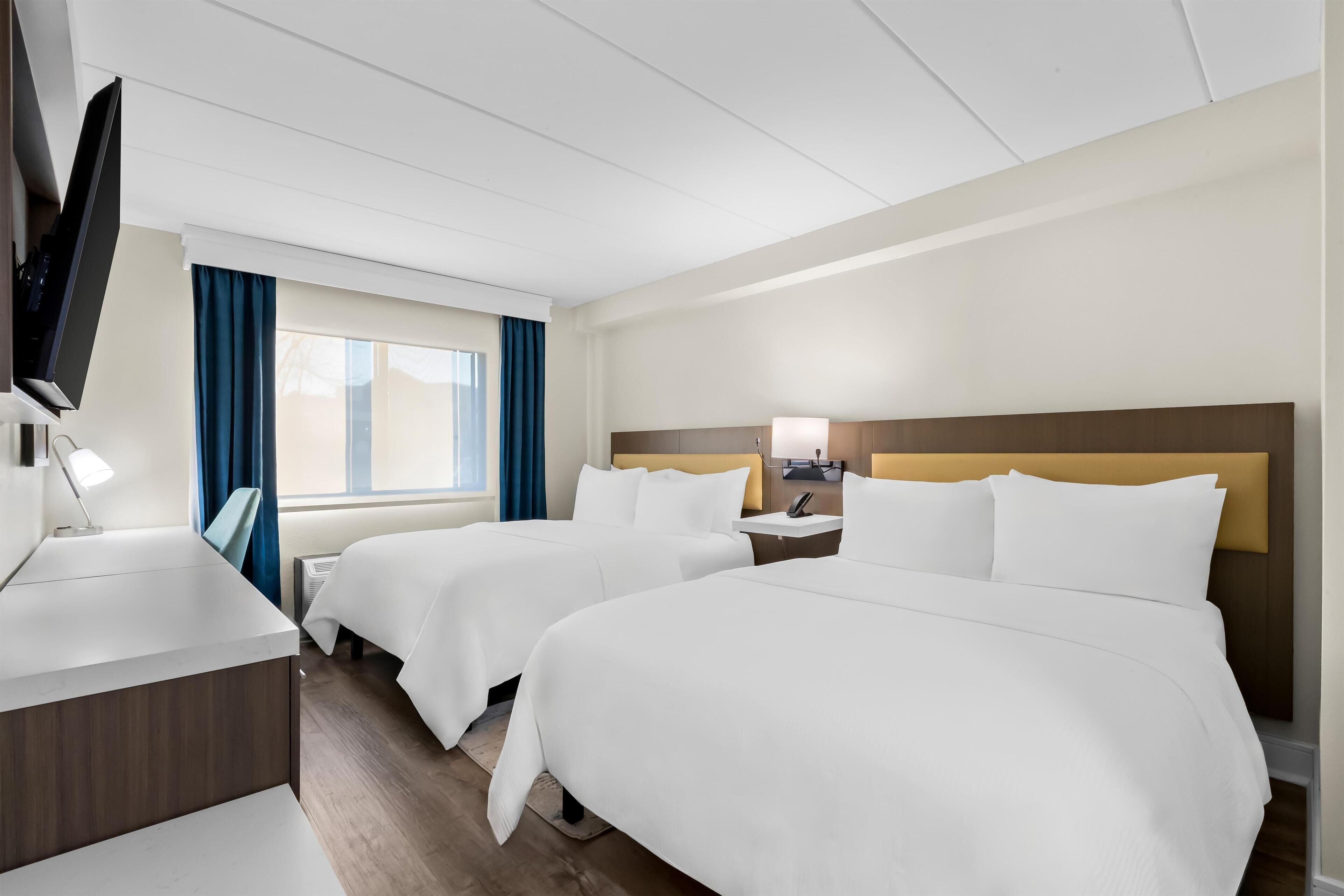 A hotel room with two beds, a desk, a TV, and a window with curtains.