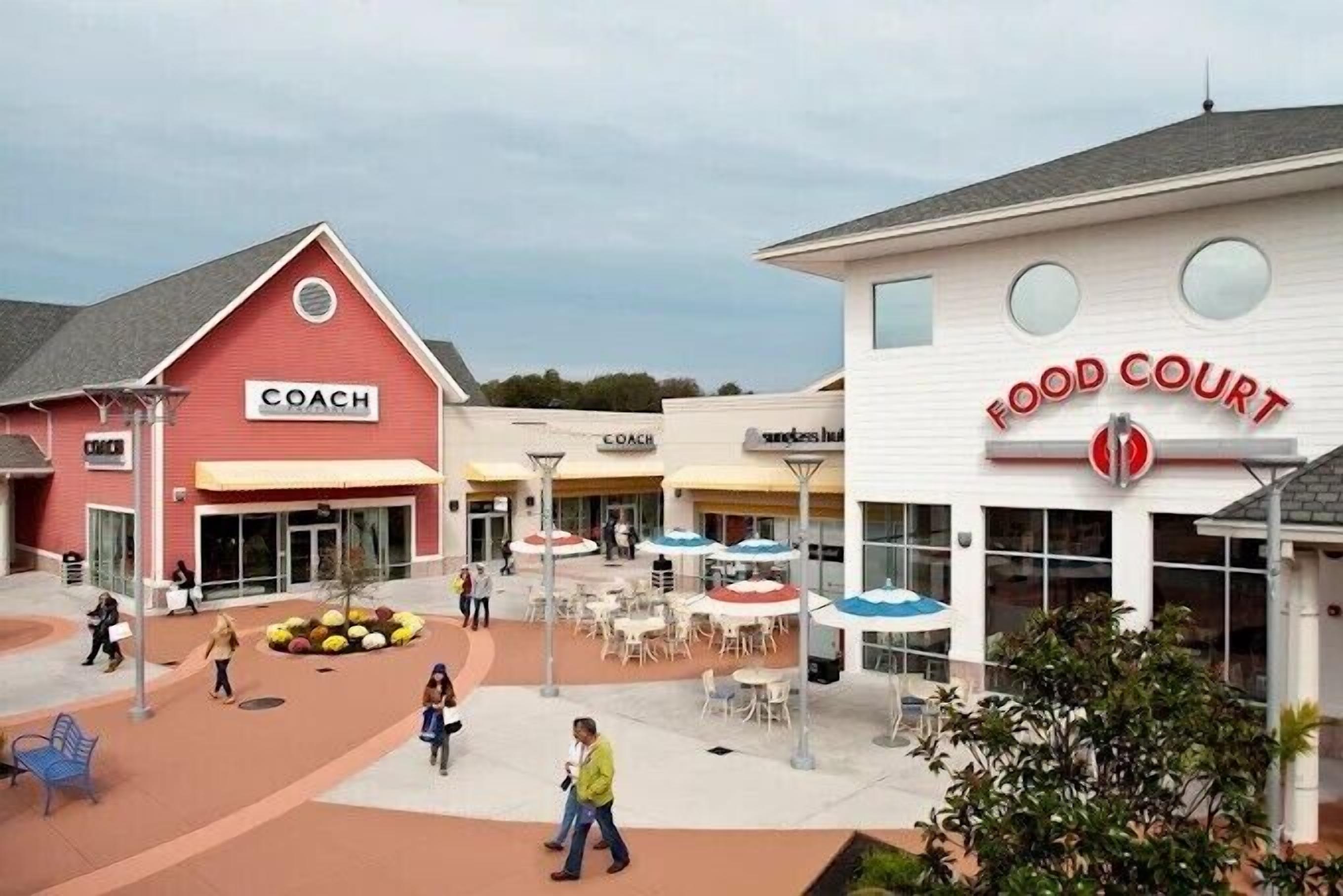 Une zone commerciale avec un bâtiment rouge portant l’inscription « COACH » et un bâtiment blanc portant l’inscription « FOOD COURT ».