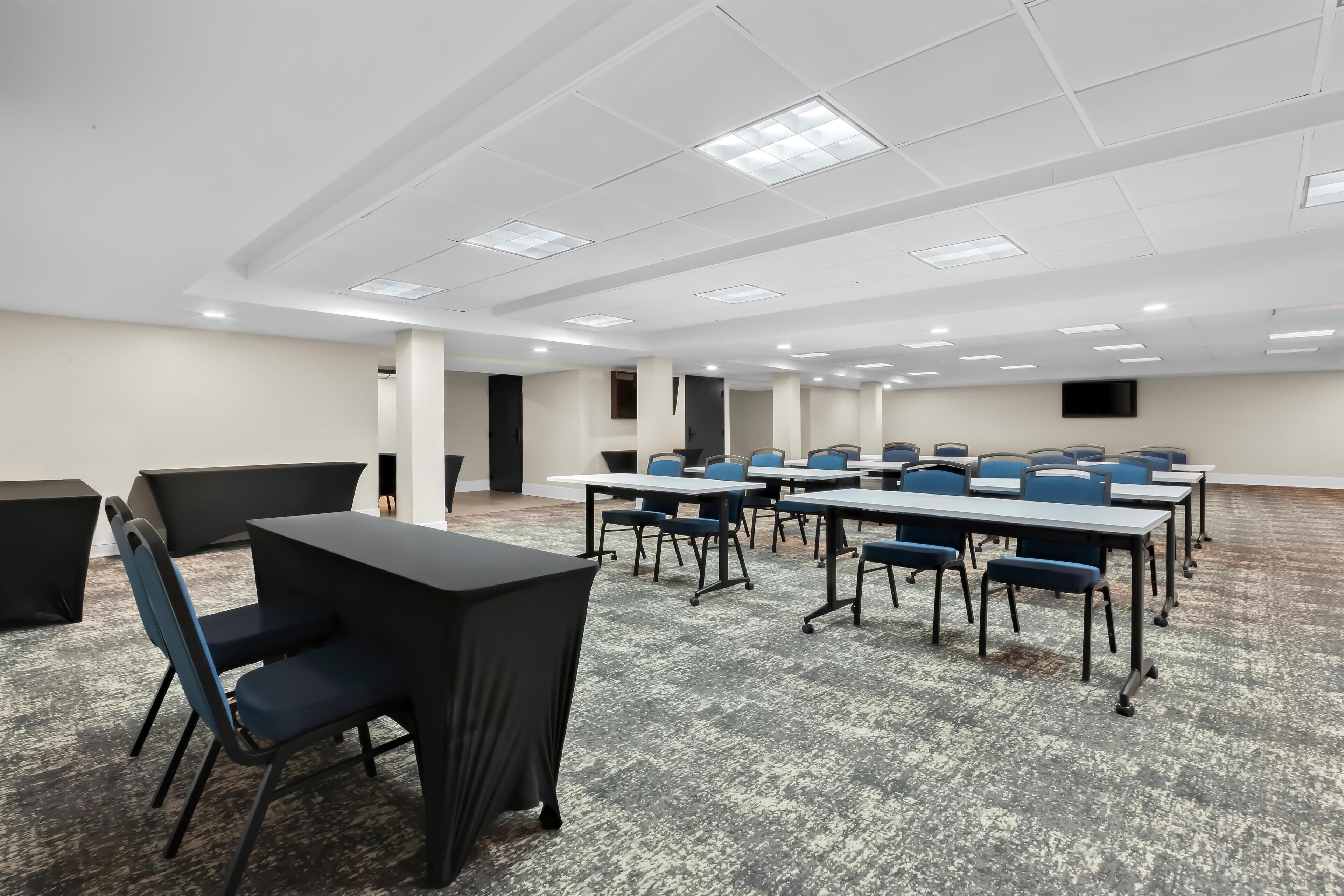Un salón de conferencias con mesas y sillas, suelo alfombrado, paredes blancas y luces en el techo.