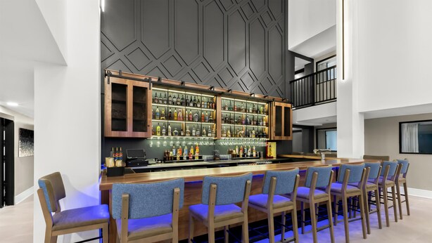Un bar moderne doté d’un long comptoir, de nombreuses étagères à bouteilles et de plusieurs chaises recouvertes d’un tissu bleu.