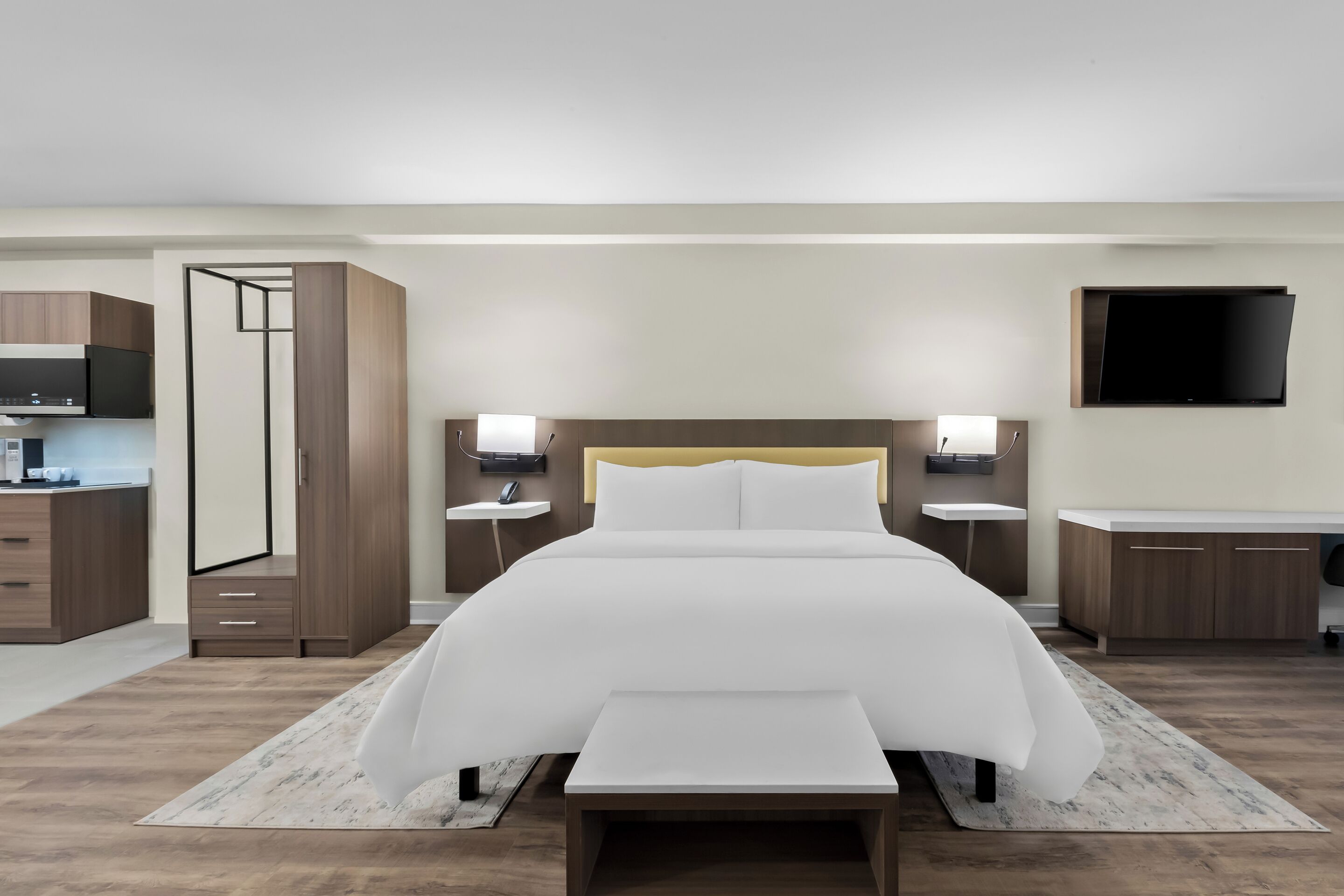 A modern hotel room with a large bed, bedside tables, a flat-screen TV, and a kitchenette.