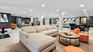 Ein großzügiger Wohnbereich mit einem großen, beigefarbenen Ecksofa, einem Holztisch und orangefarbenen Hocker. Dunkelblaue Sessel und ein kleiner Tisch mit Schachbrett vervollständigen das Bild. Das Zimmer hat ein modernes Design, ein großes Fenster und einen Blick auf die Stadt.