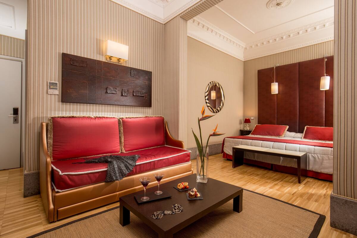 A hotel room with a red sofa, a bed with red bedding, a coffee table with a vase and two wine glasses, and a wall with a large artwork and a mirror.